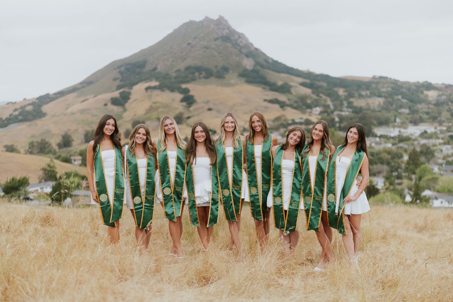 Large group in white dresses poses with raised arms in front of a mountain landscape.