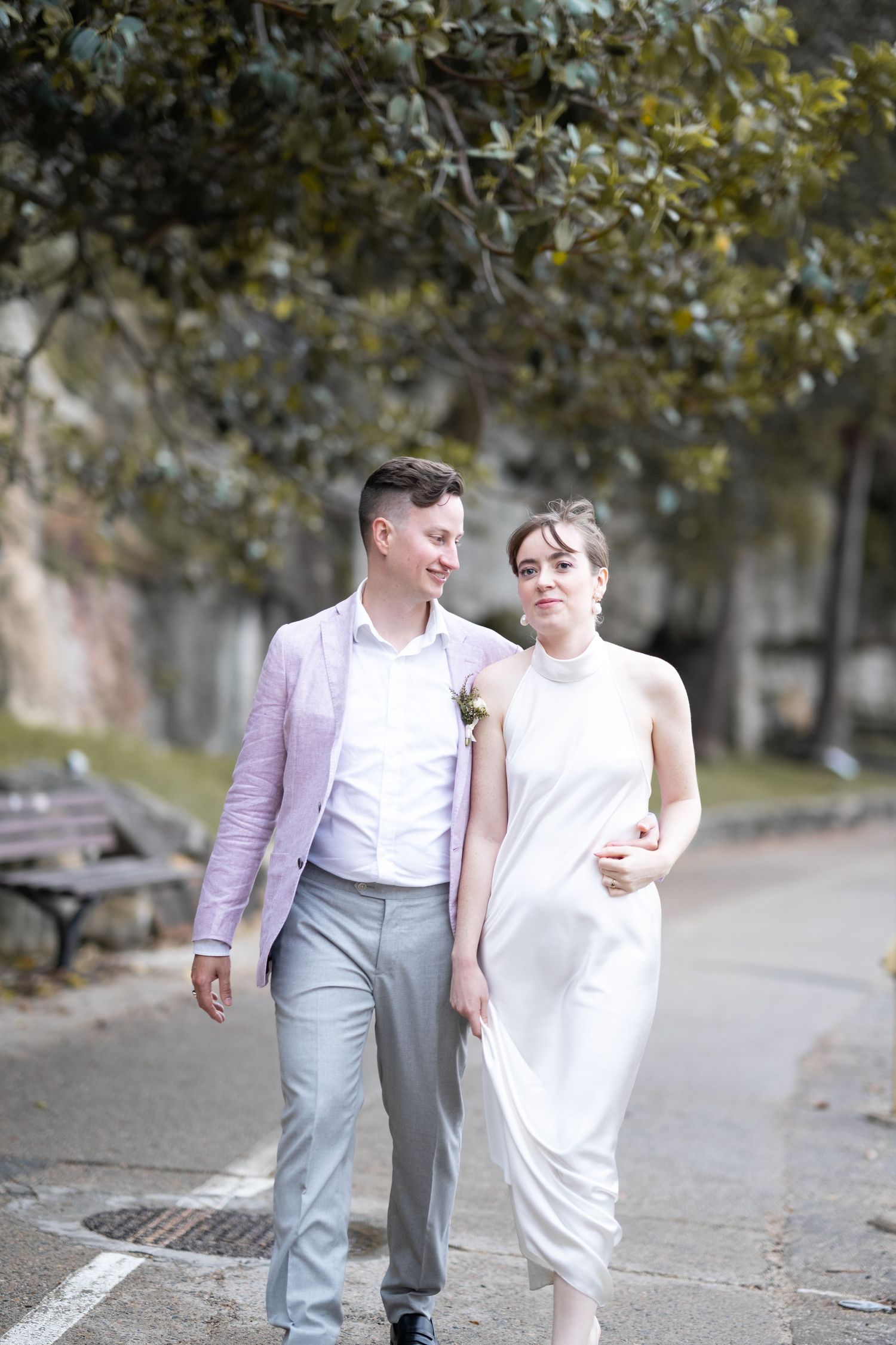 A bride in white dress and groom in light blazer walk together on a tree-lined path.