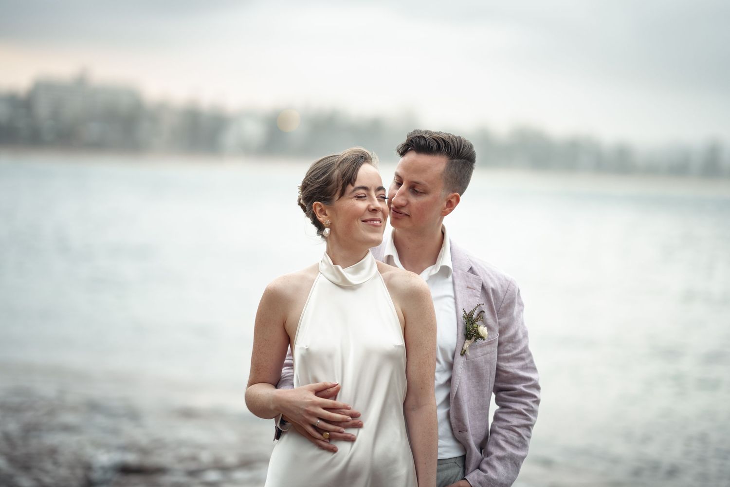 A couple shares a tender moment by a serene waterfront in formal attire.