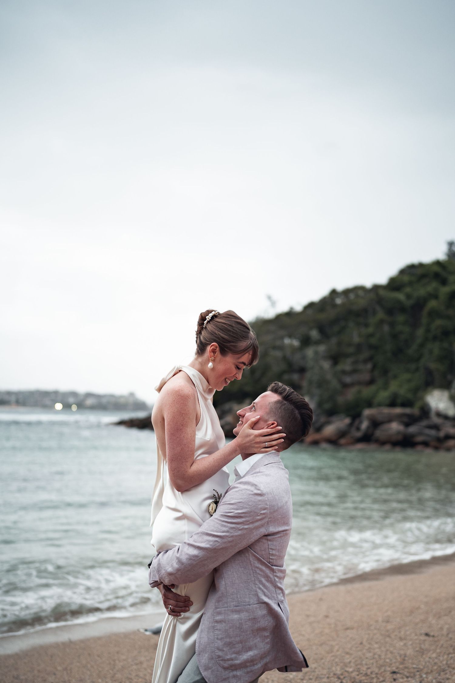 A couple in wedding attire embraces passionately on a beach with rocky cliffs in background.