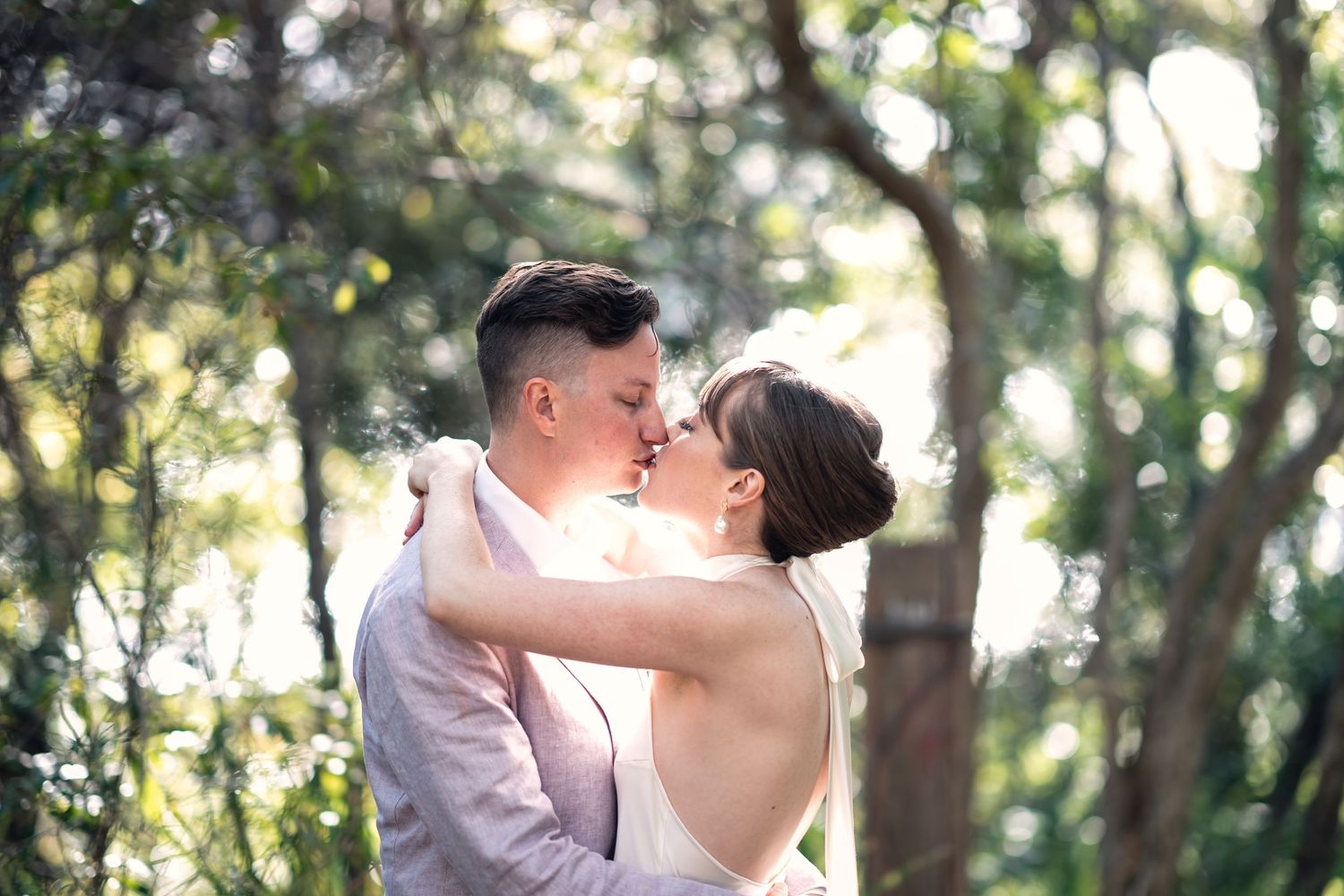 A romantic wedding couple embraces in a sun-dappled forest setting.
