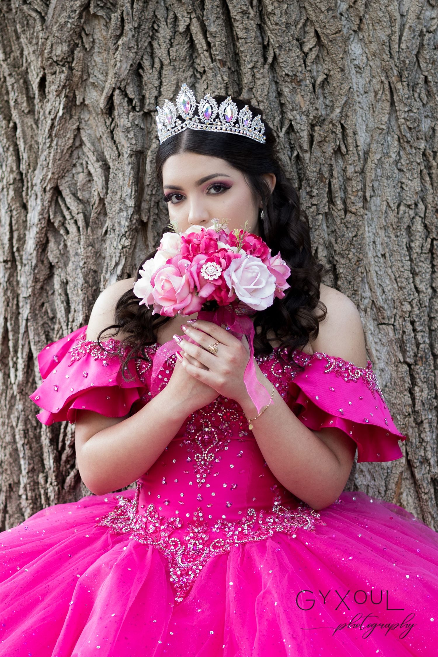 Hot pink quinceañera dress with crystal embellishments and matching floral bouquet against tree trunk.