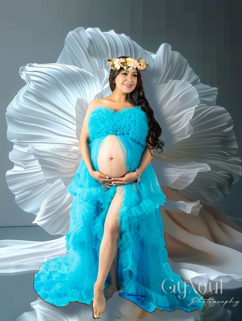 A maternity photo showcases a dramatic turquoise gown against a white pleated backdrop.