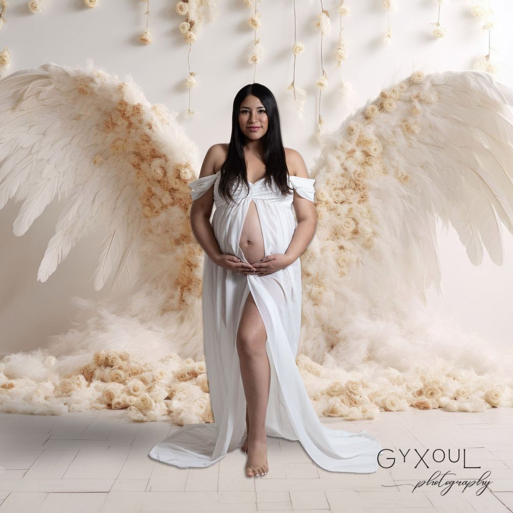 Dramatic maternity photo with large angel wings and flowing white gown against cream backdrop.