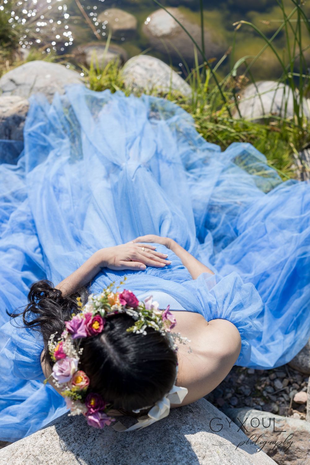 A dreamy maternity photo features a flowing blue dress and floral crown by a rocky stream.