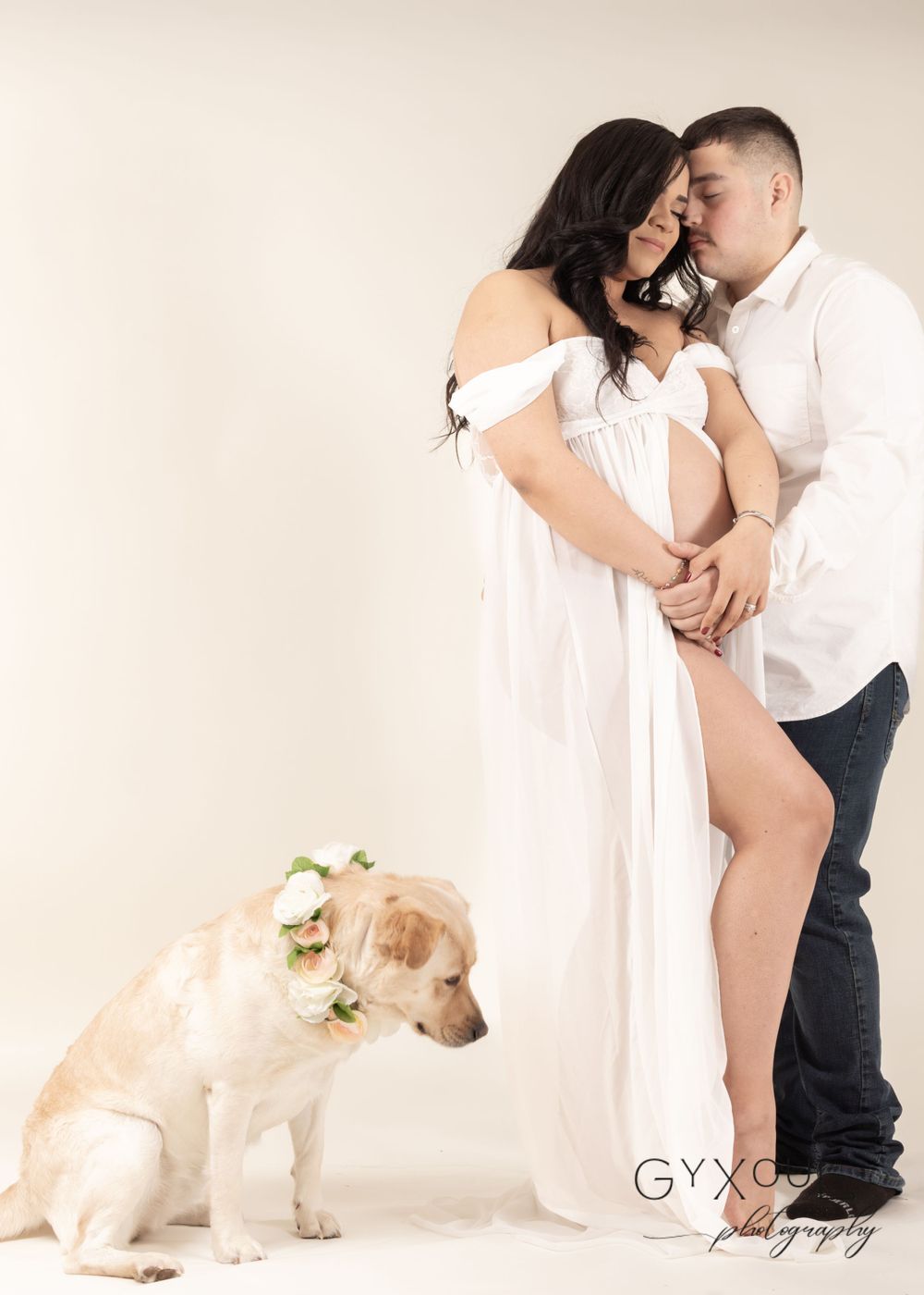 Intimate maternity portrait with couple and dog in white dress against neutral background.