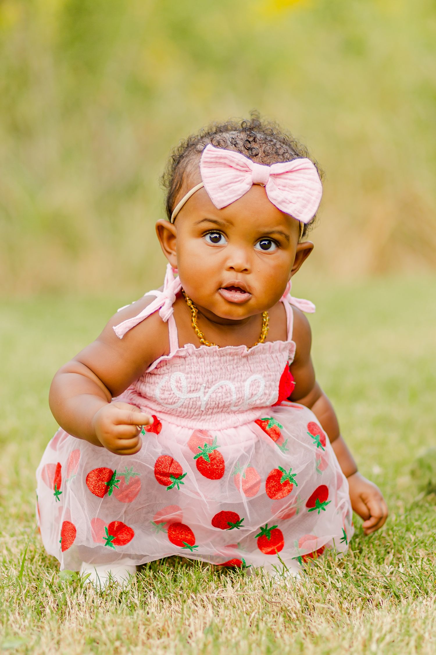 A baby wearing a pink bow and strawberry dress sits in grass during a photo session.