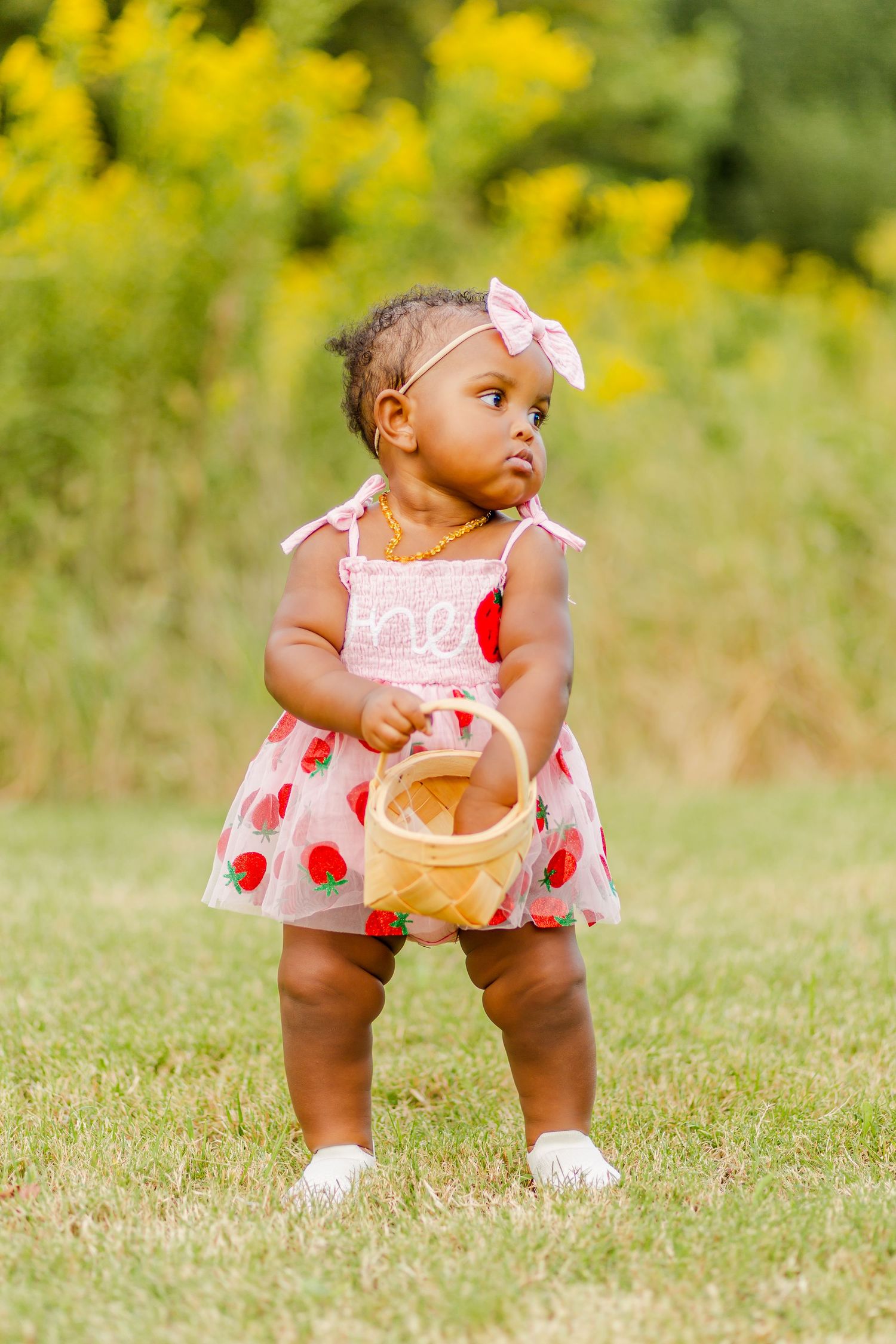 A baby in a strawberry dress and bow headband holds a wicker basket outdoors.