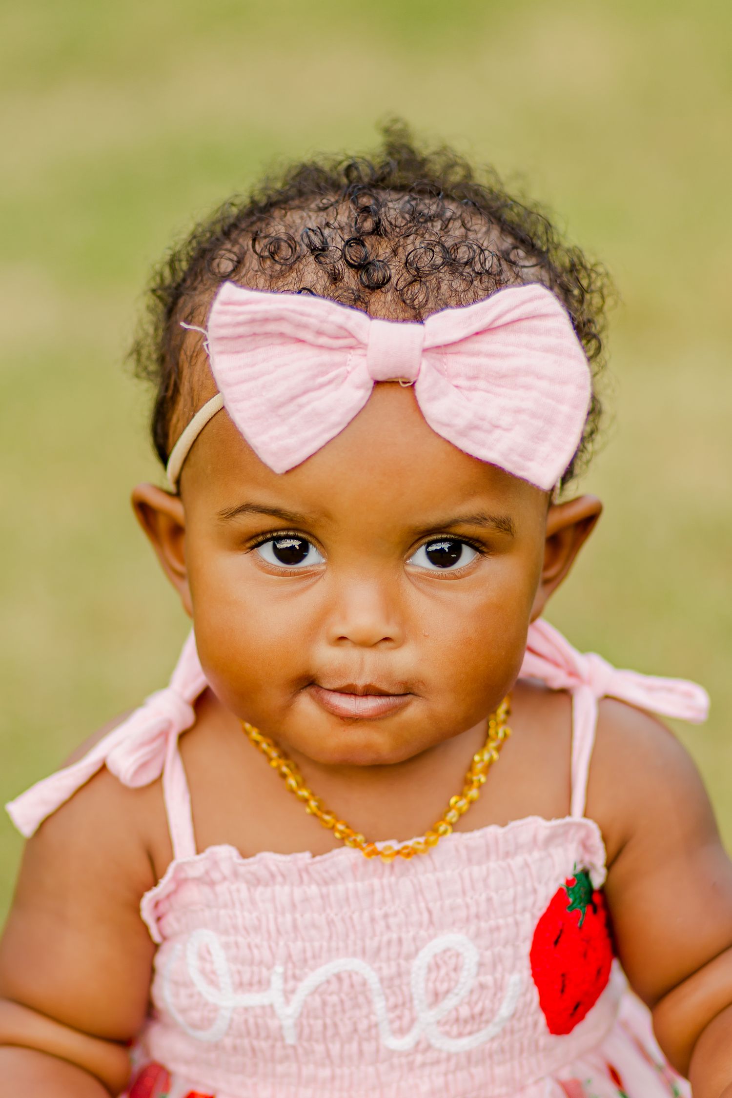 A close up portrait of a baby wearing a pink bow headband and gold necklace.