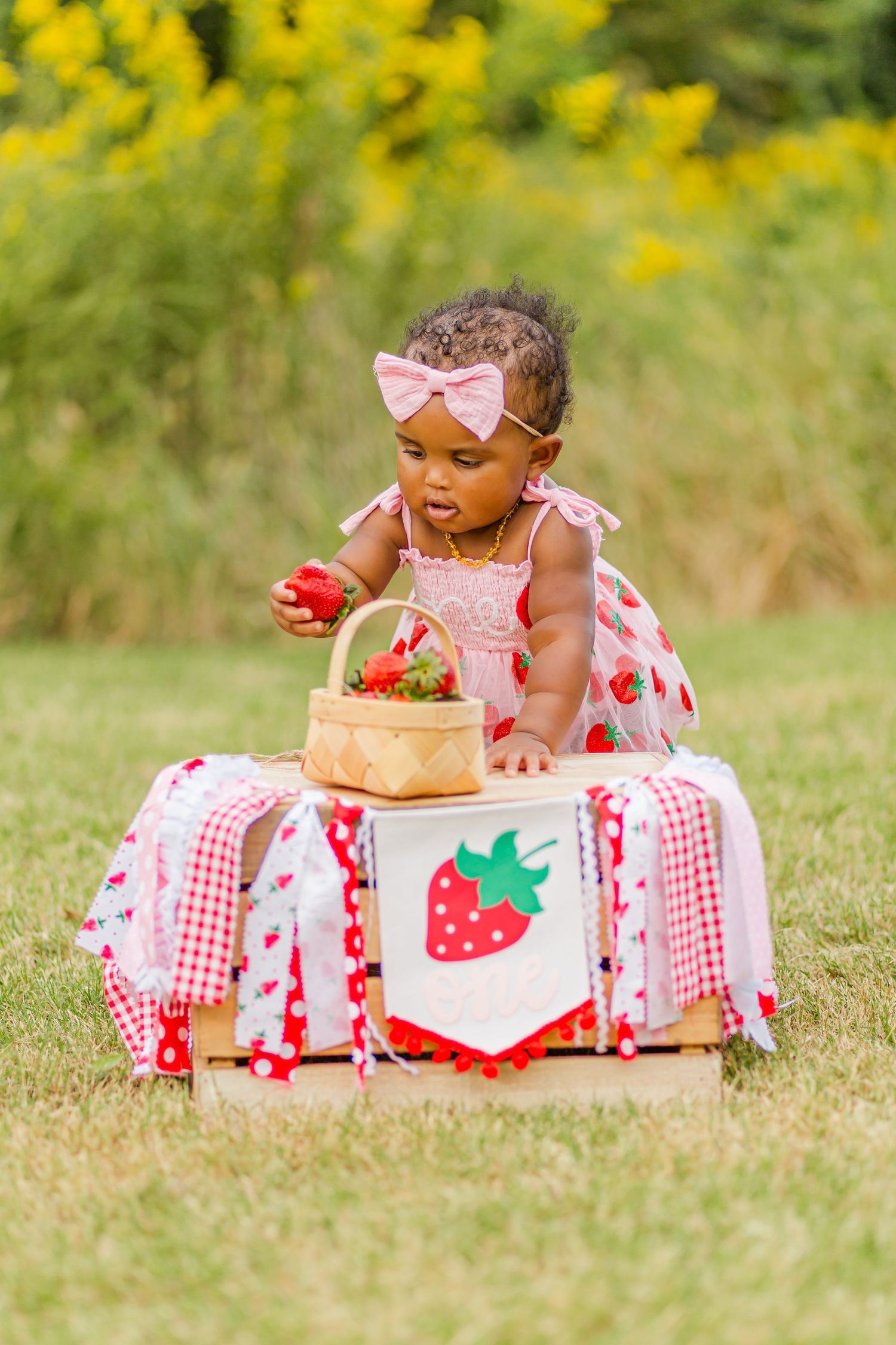A baby in strawberry-themed attire explores decorative props during an outdoor photo session.