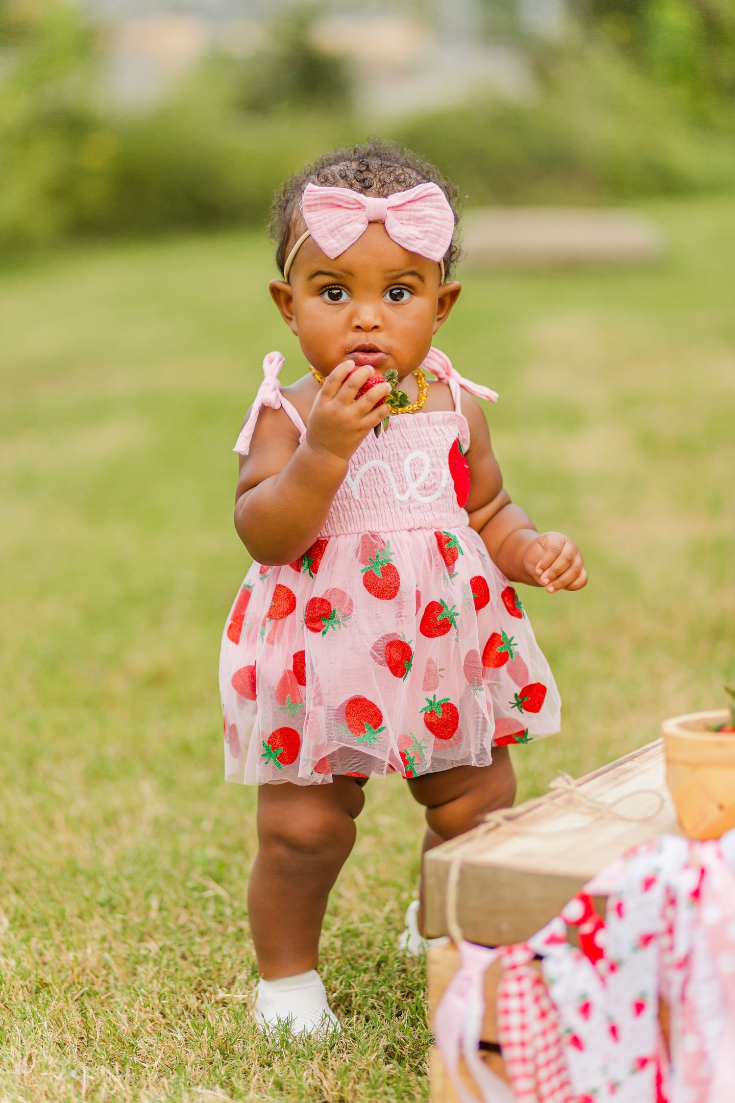 A baby in a pink strawberry dress reaches for something during an outdoor photo session.