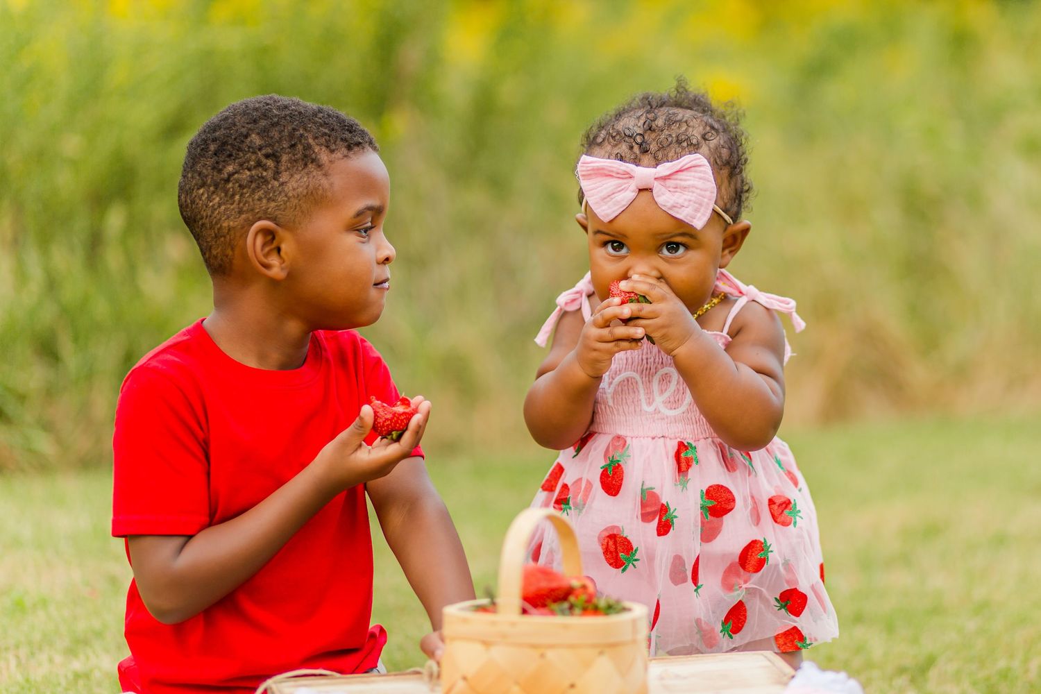 Two children share strawberries from a basket during an outdoor picnic.
