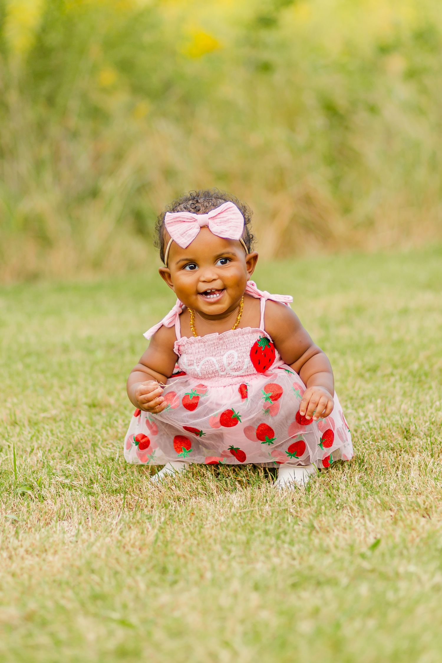 Happy baby sitting in grass wearing pink strawberry dress and matching hair bow.