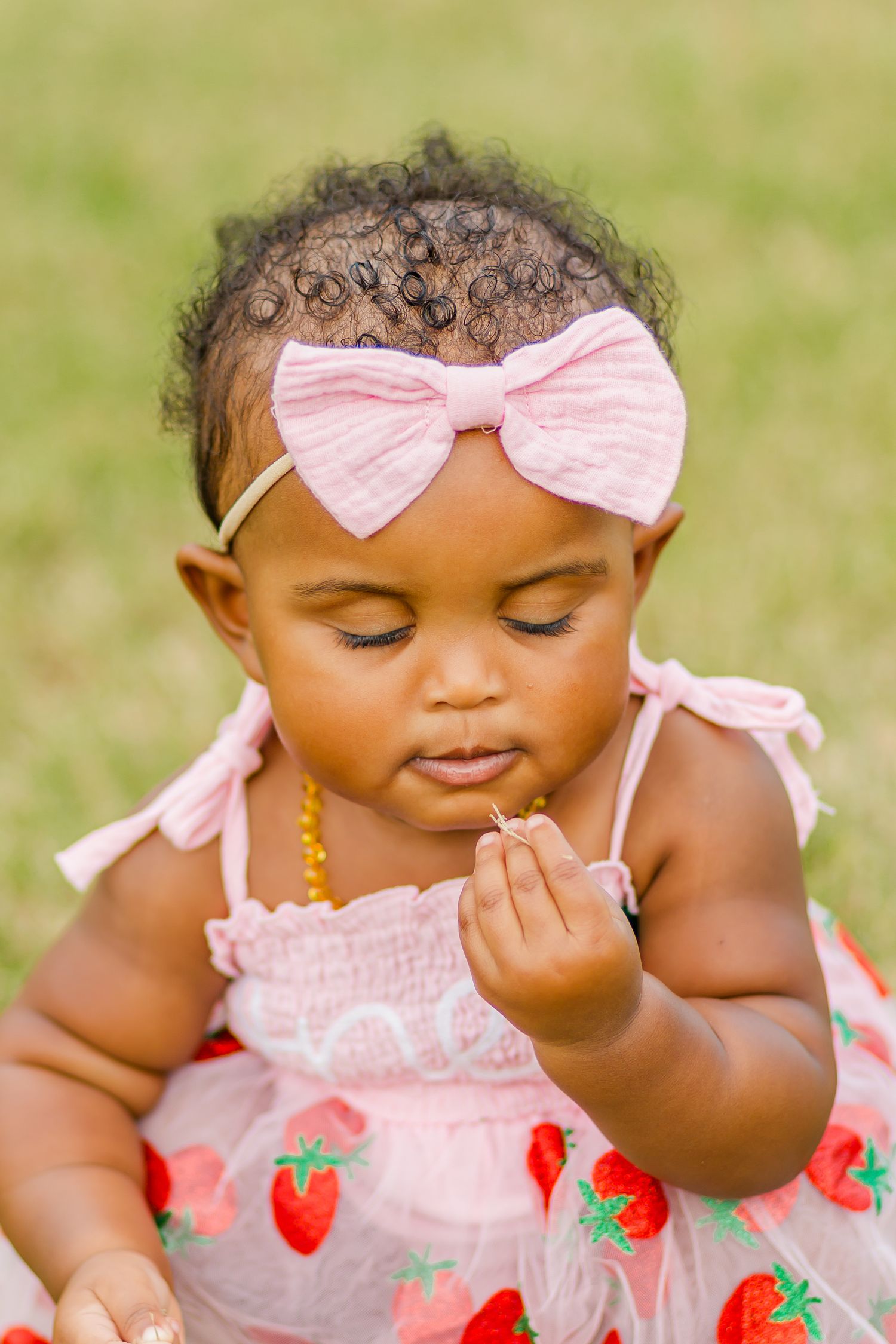 Baby in strawberry print dress with pink bow looking down thoughtfully.