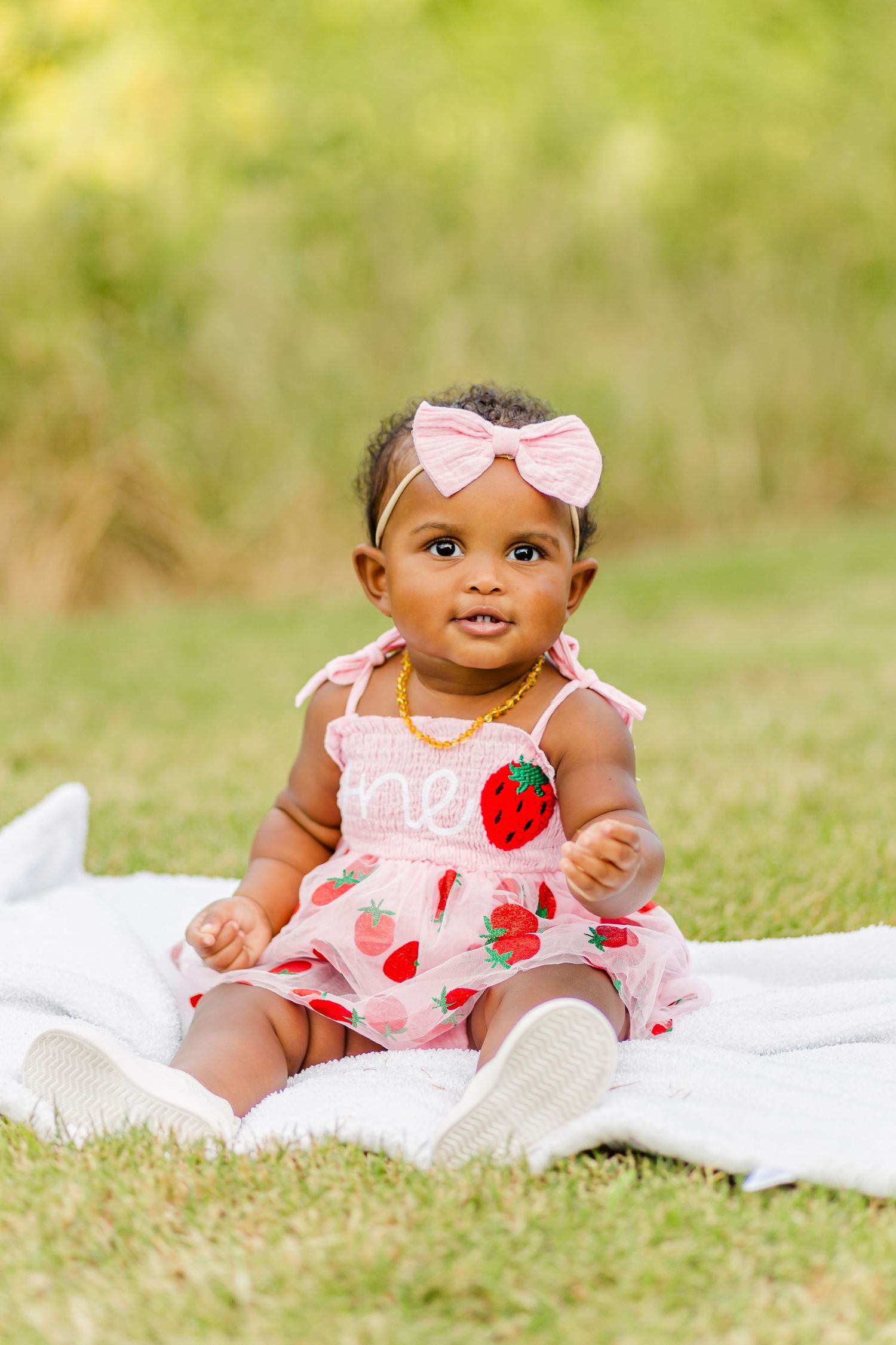 A baby in a pink and white strawberry-print dress sits on a blanket outdoors.