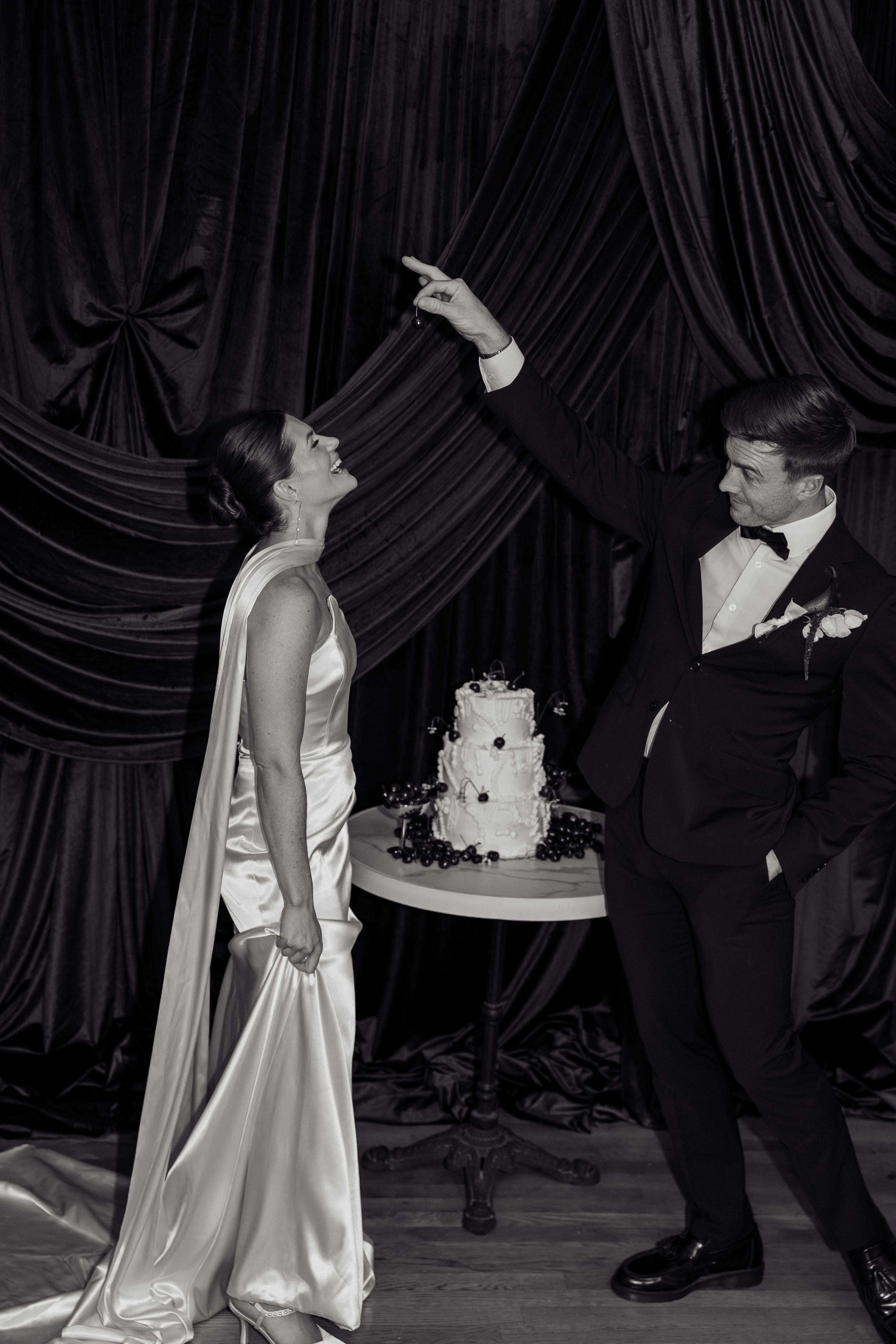 Black and white photo of bride and groom laughing beside wedding cake, groom pointing upward in tuxedo.
