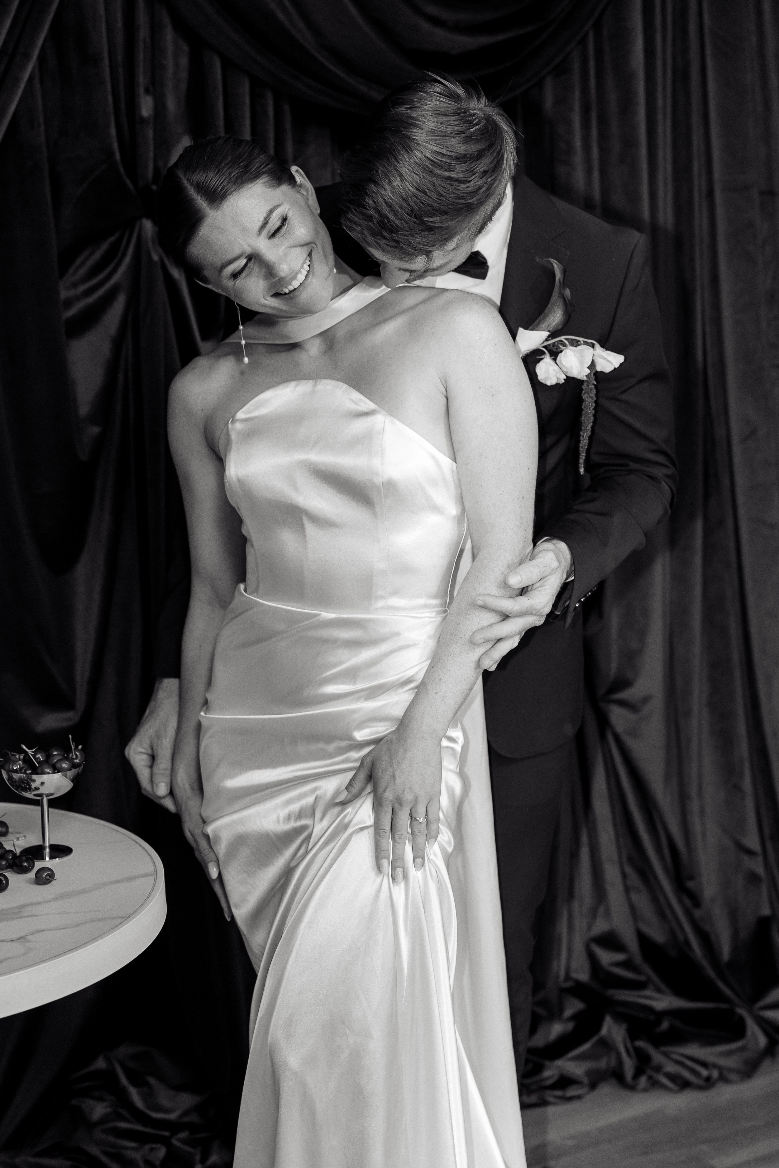 Black and white photo of elegant bride in satin gown embraced by groom in tuxedo, sharing romantic moment.