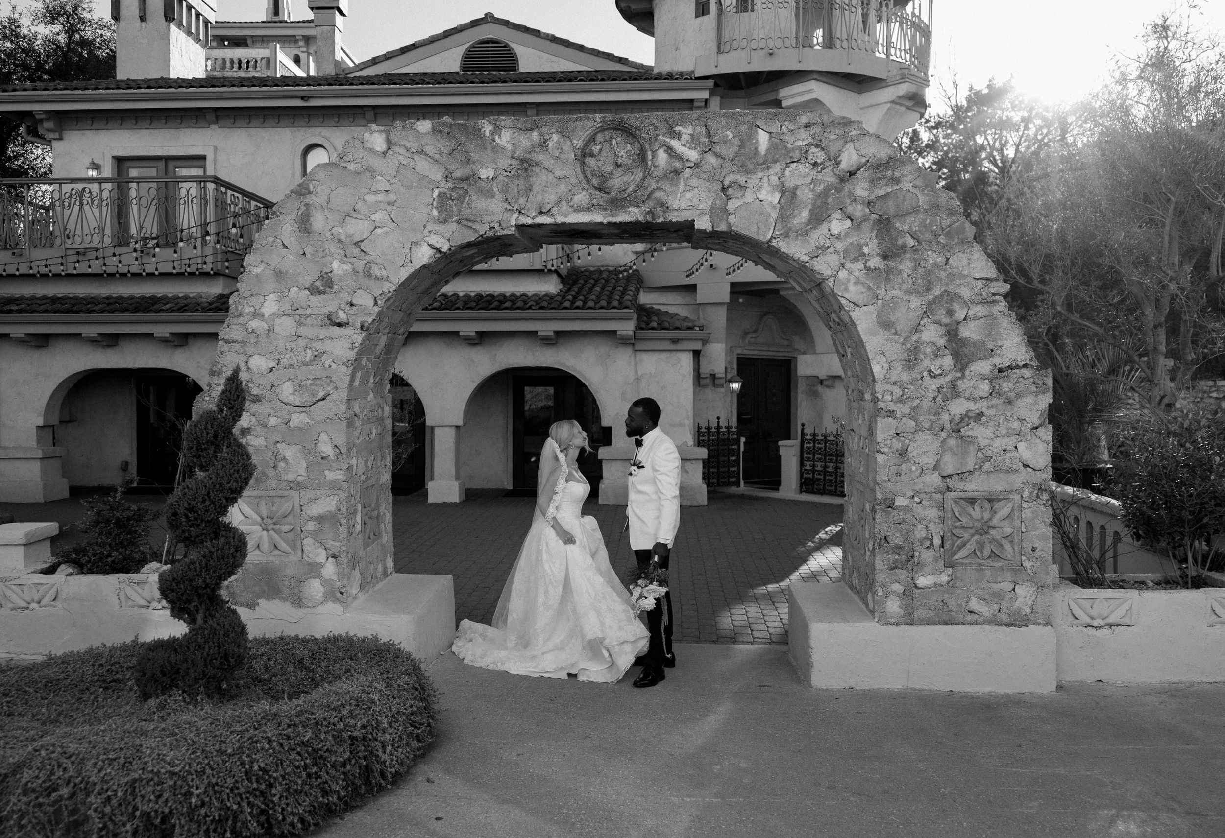 A black and white wedding moment captured under a rustic stone archway at an elegant venue.