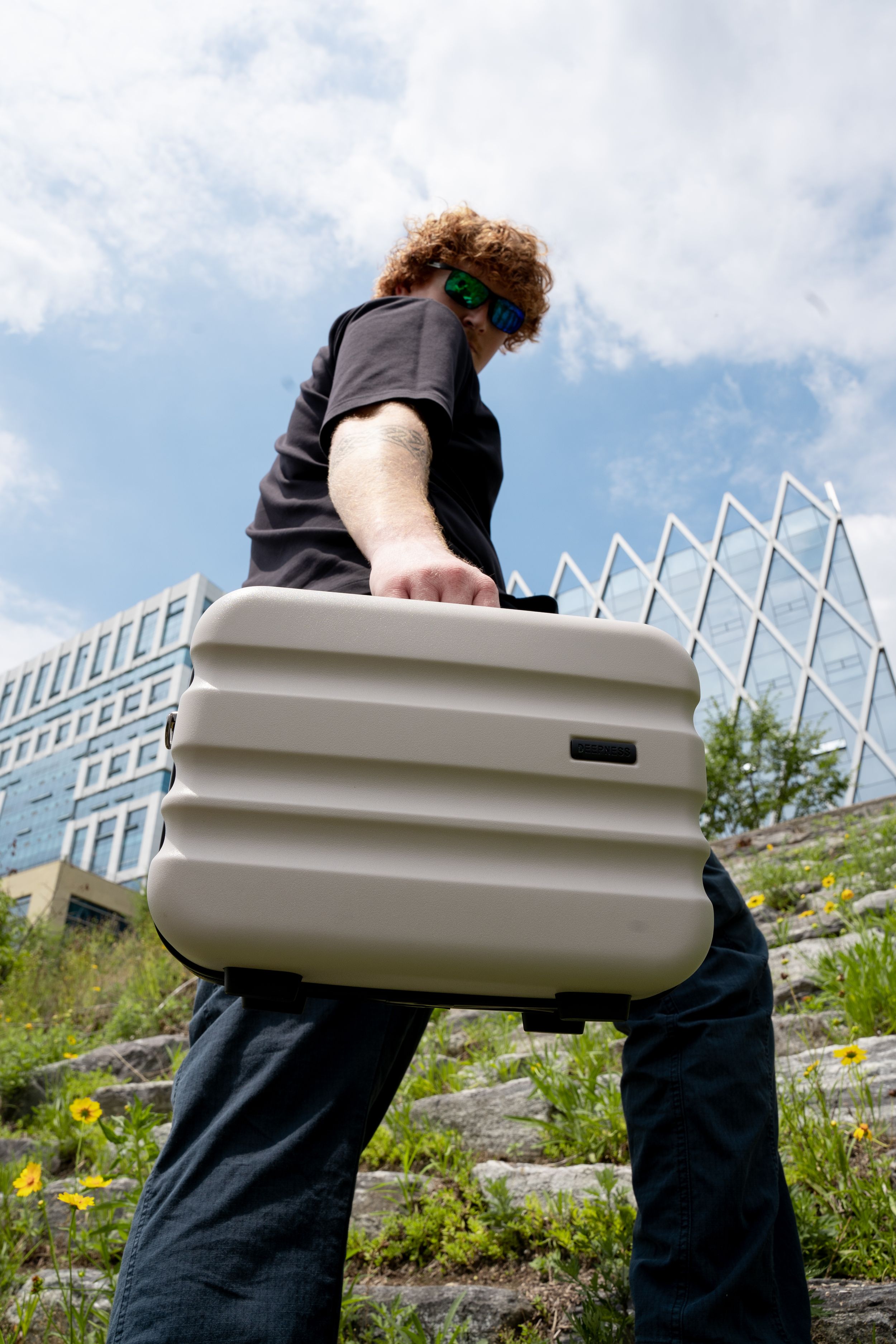 Person carrying beige hardshell luggage case in front of modern building architecture.