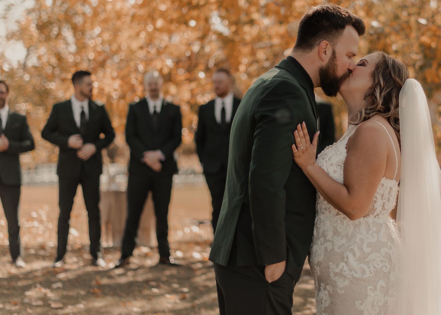 Bride and groom share a first look kiss with groomsmen in the background, set against a fall foliage..