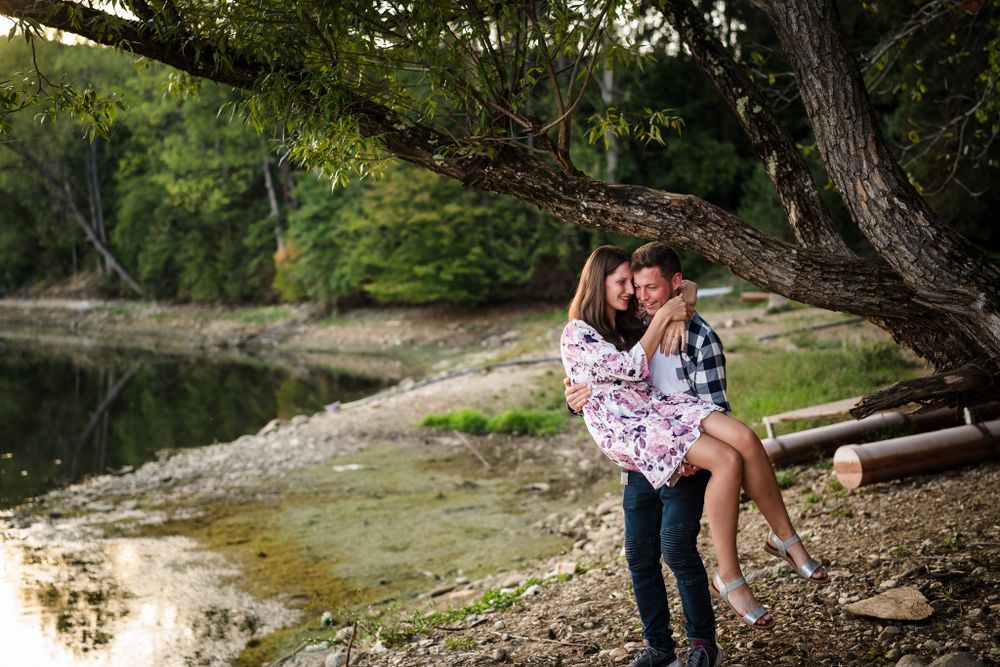 A couple embraces playfully by a scenic lake with trees and rocks in the background.