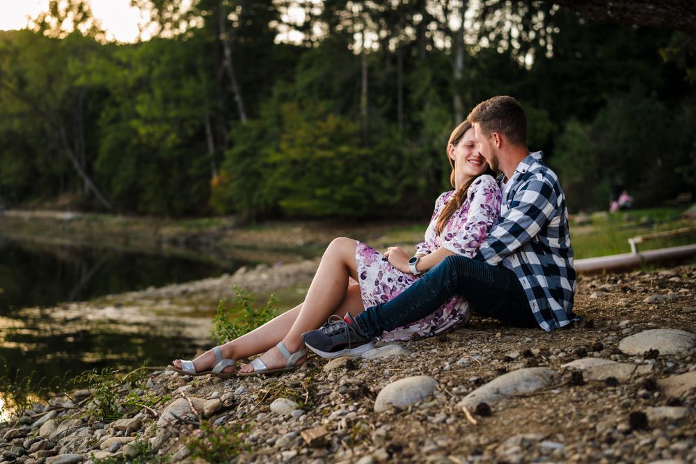 A couple sits together on rocky shoreline during golden hour beside a tranquil lake.