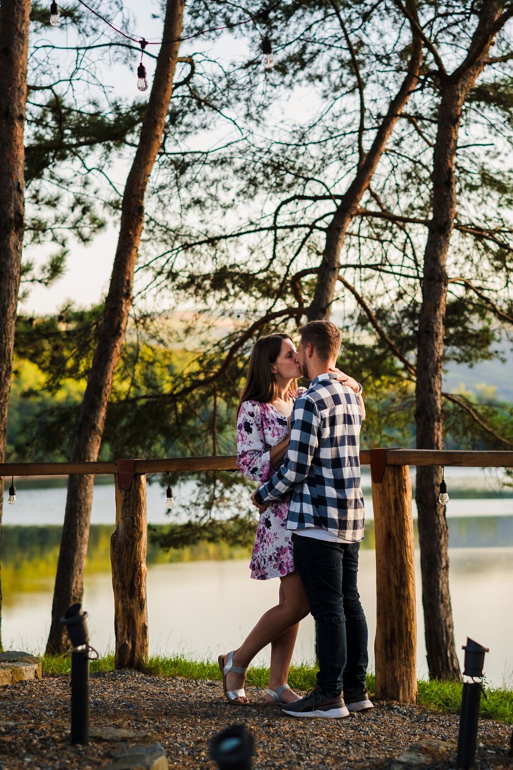 A romantic scene on a lakeside deck with pine trees framing the couple at sunset.