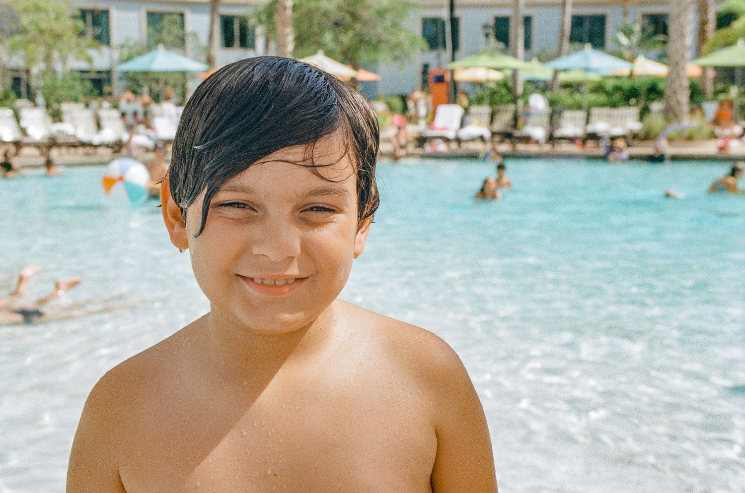 A child smiles at a pool with other swimmers visible in the background.