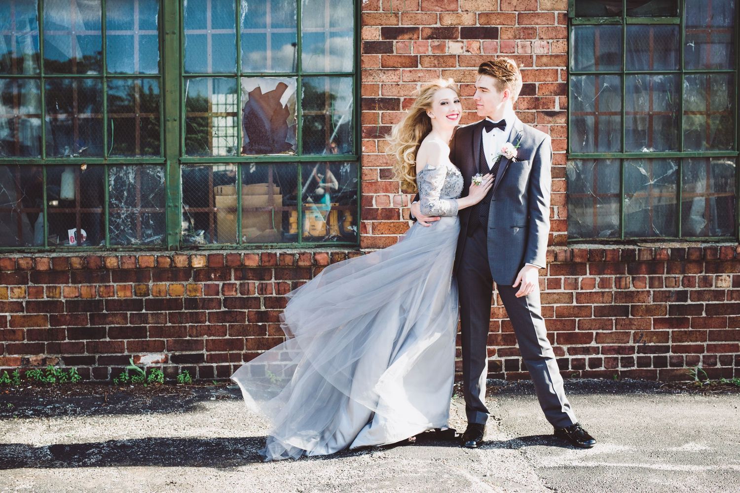 A couple poses against a brick wall with large windows as her flowing dress catches the wind.