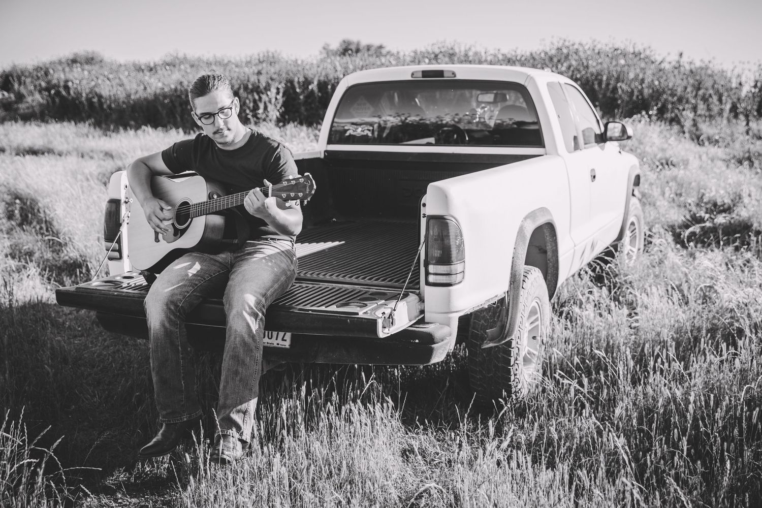 A black and white photo of someone playing guitar while sitting in a pickup truck bed.