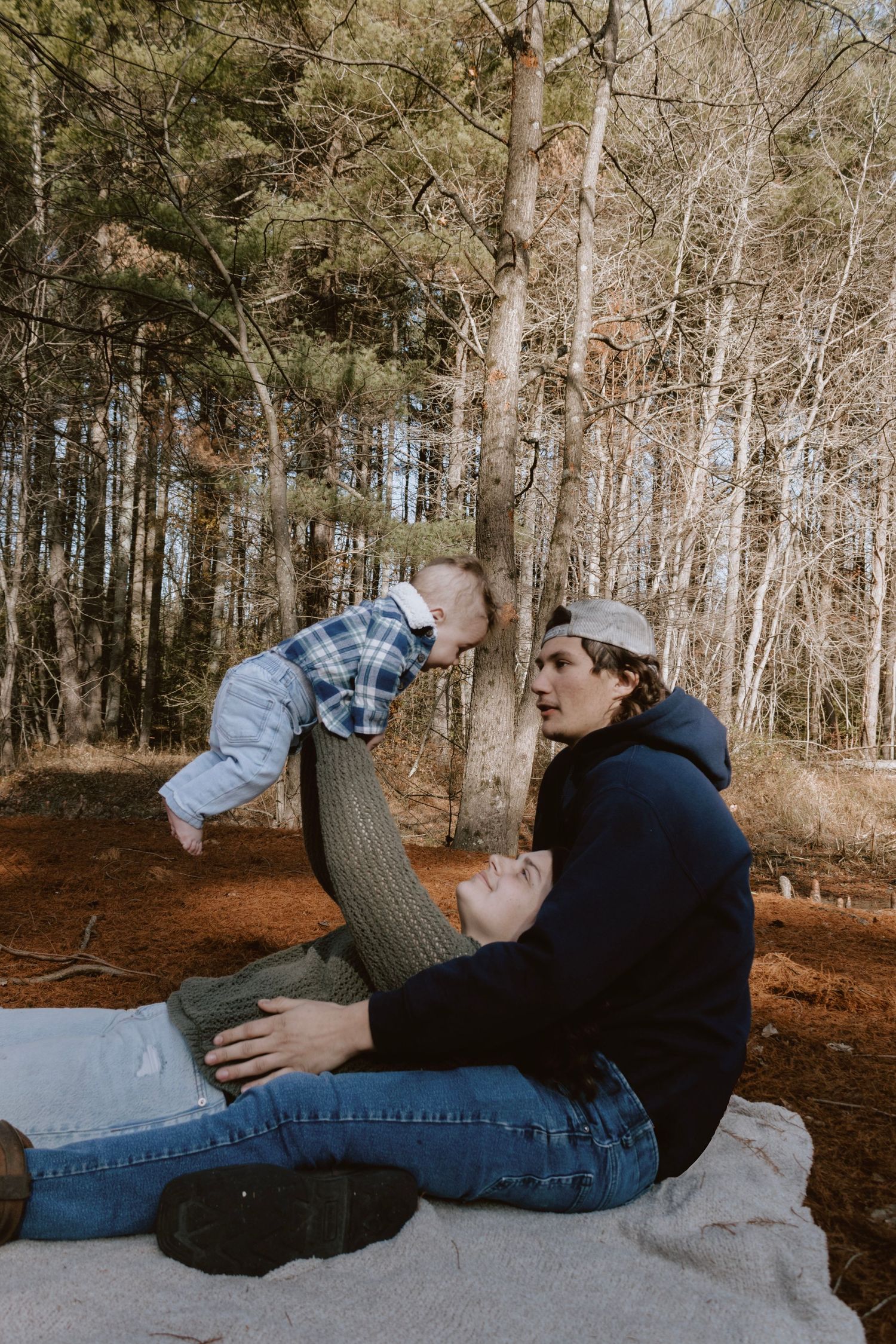 Parent and baby share playful moment on blanket in wooded setting.