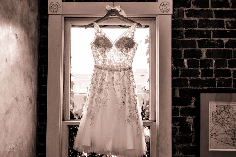 A delicate white lace wedding dress with intricate beading hangs in a doorway against exposed brick wall.