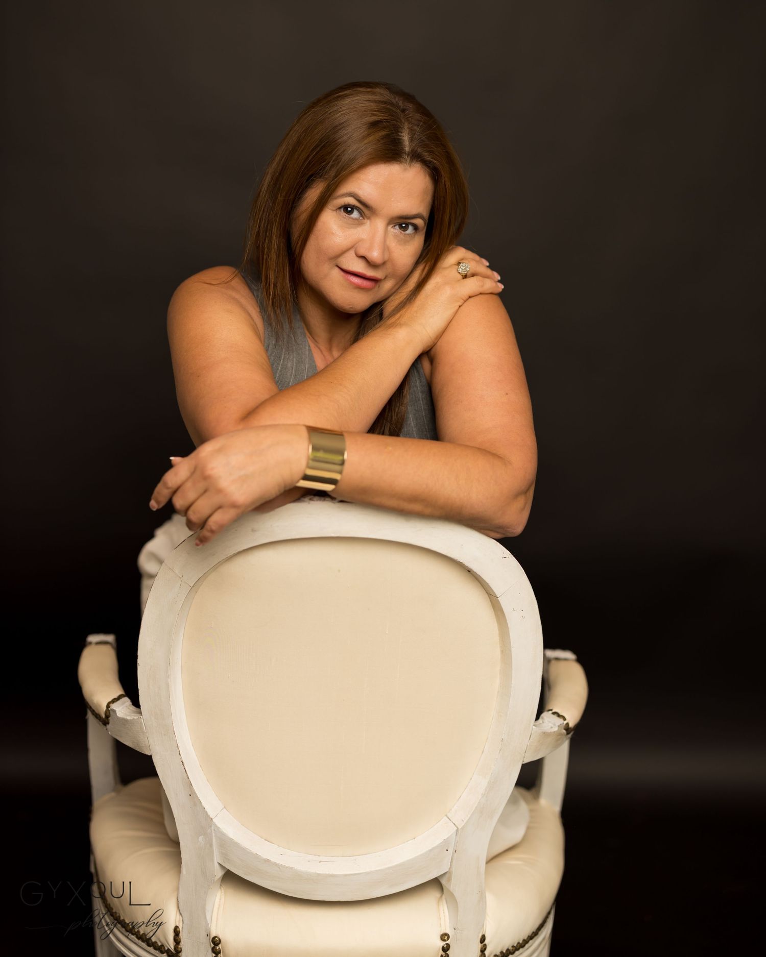 Professional portrait in studio setting with person leaning on white chair against dark background.