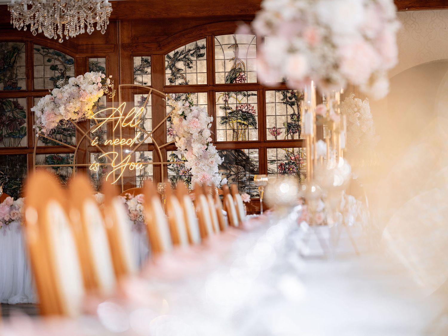 Elegant wedding reception table setup with crystal chandelier and soft floral centerpieces in a sunlit room.
