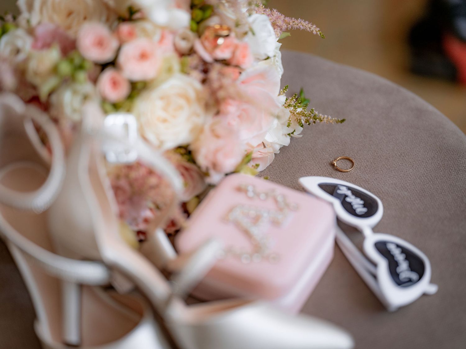 Wedding accessories with pink floral bouquet, white heels, and sunglasses on grey surface.
