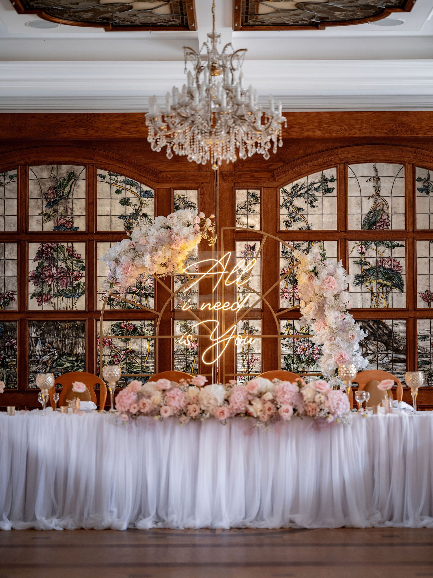 Elegant wedding reception setup with crystal chandelier and floral arrangements against stained glass windows.