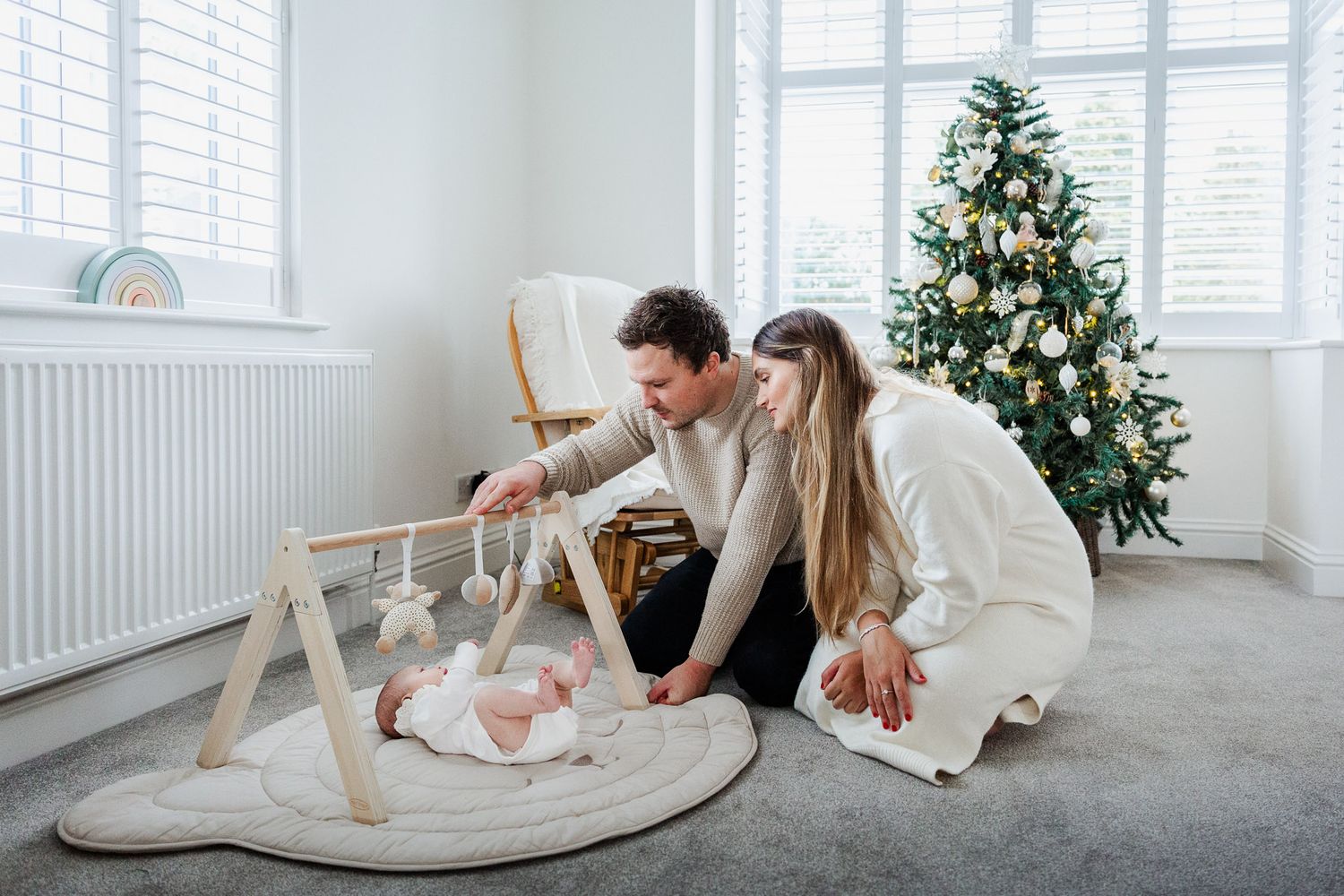 A mum and dad play with their newborn baby in front of the Christmas tree at home. Bristol family photographer