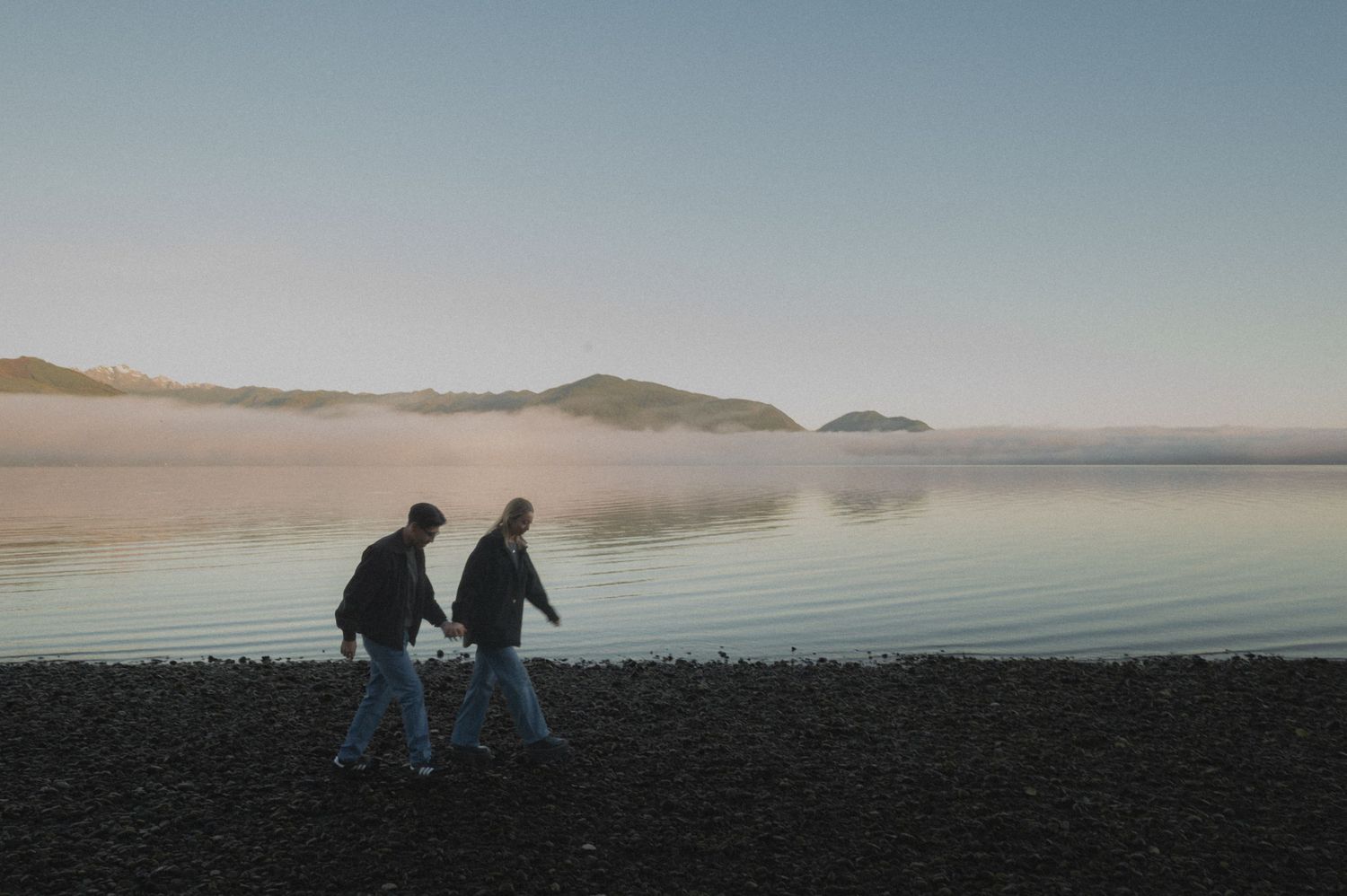Two people stroll along a misty beach shoreline at sunset.
