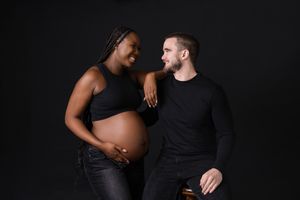 Black and white maternity photo of an expecting couple sharing a joyful moment against dark background.