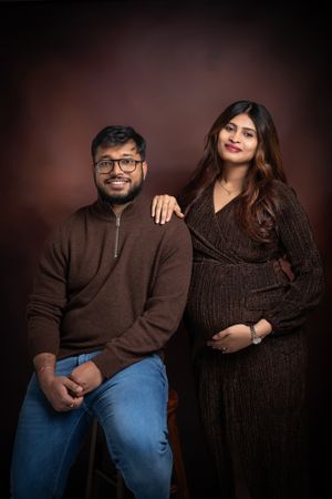 Studio portrait of expecting couple in brown sweaters against dark background.