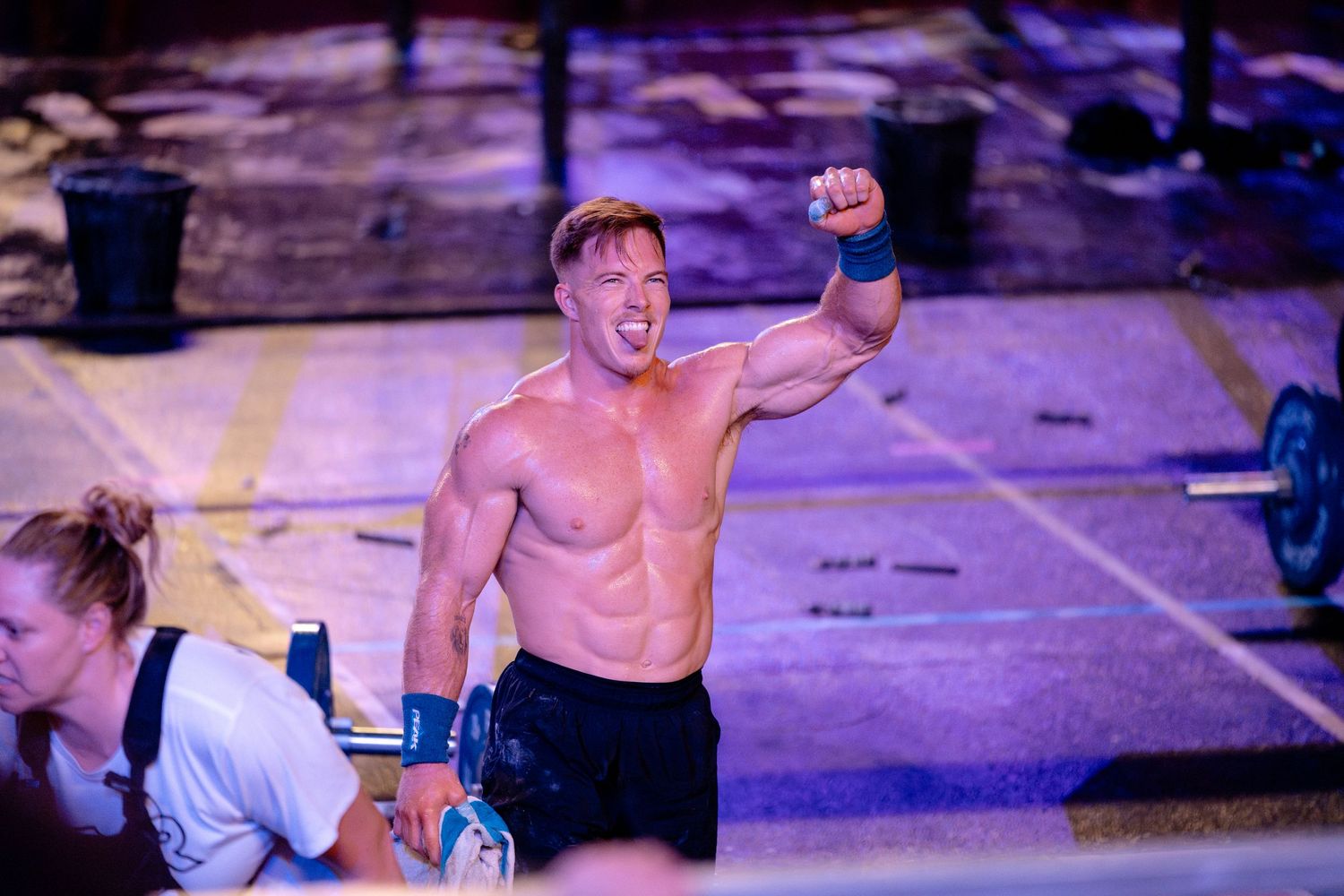 Shirtless athlete celebrates victory at outdoor CrossFit competition with purple lighting.
