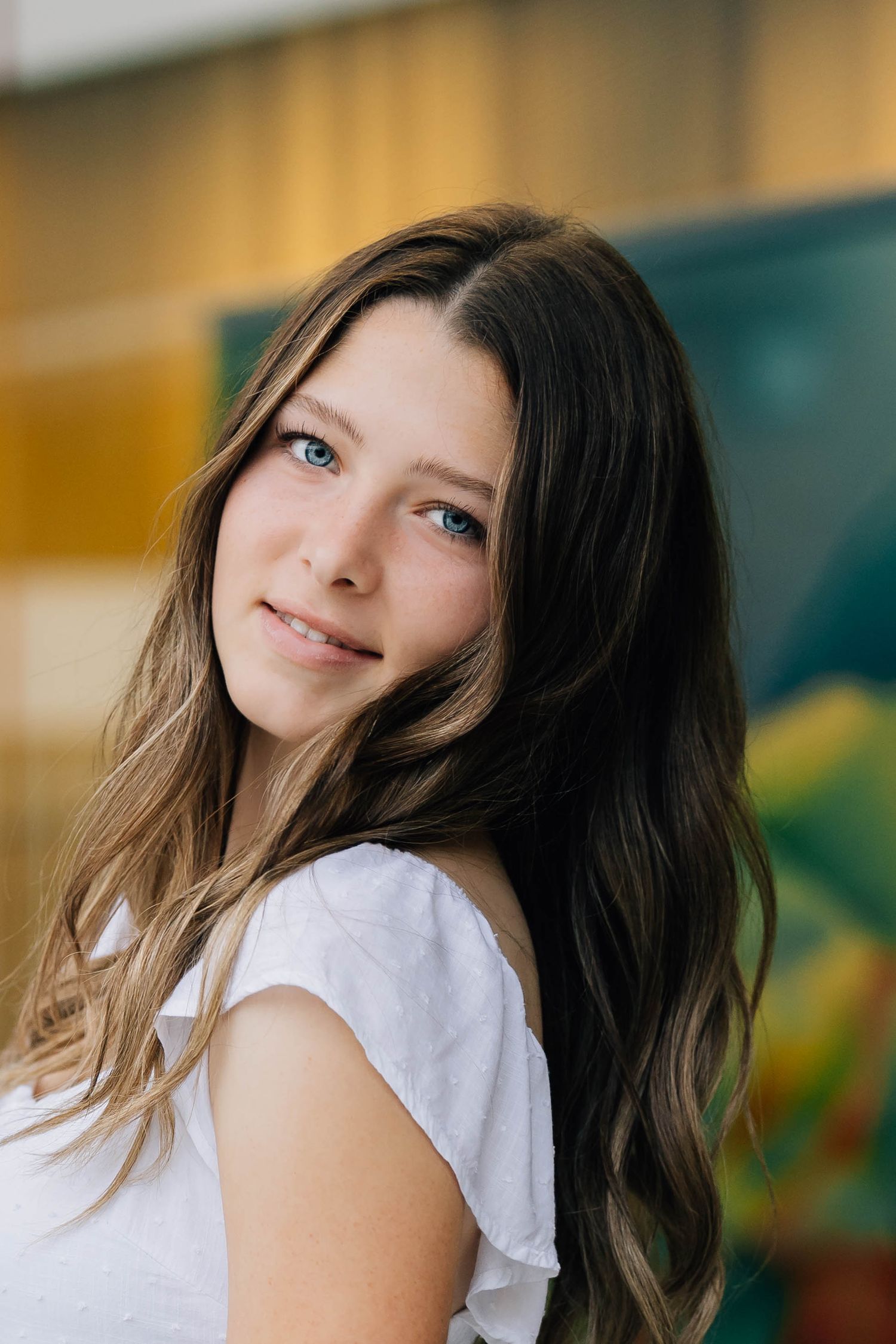Teen girl with long brown hair and blue eyes smiling in a white top indoors.