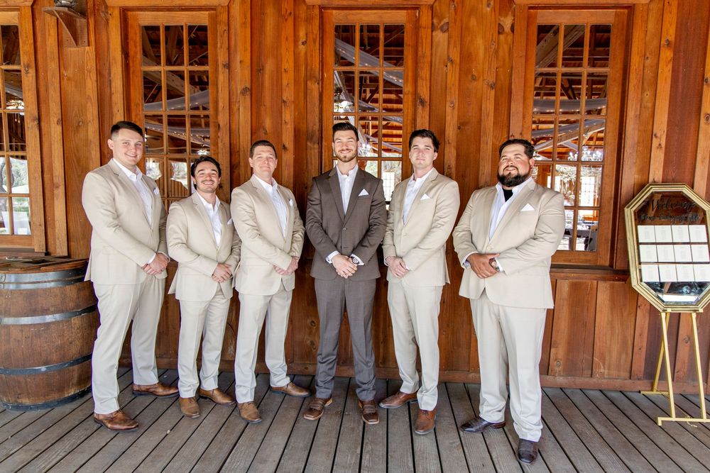 Wedding party in light beige suits poses with groom in charcoal grey on rustic wooden porch with barrels.