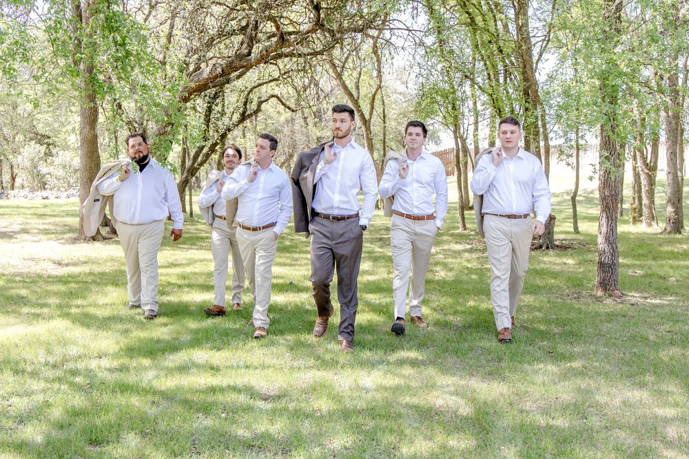 Five groomsmen in matching light-colored attire walk together through a sunlit wooded area on a bright summer day.