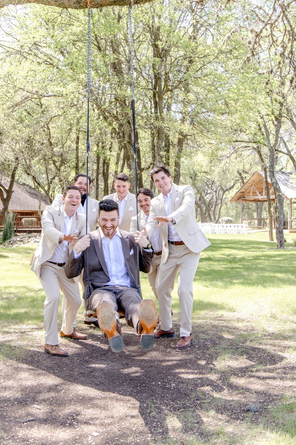 A group in light colored attire enjoys a playful moment on a rustic rope swing in a sunlit wooded outdoor setting.
