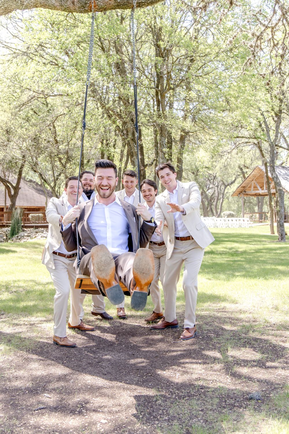 Group wearing khaki suits and white shirts playfully lift someone on a swing outdoors at a rustic venue.