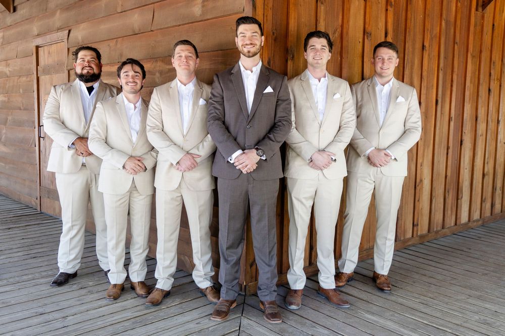 Groom in gray suit poses with groomsmen in light beige suits standing in front of wooden wall at wedding venue.