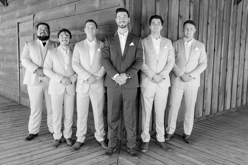 Black and white photo of a groom in dark suit standing with groomsmen in light suits on a wooden deck.