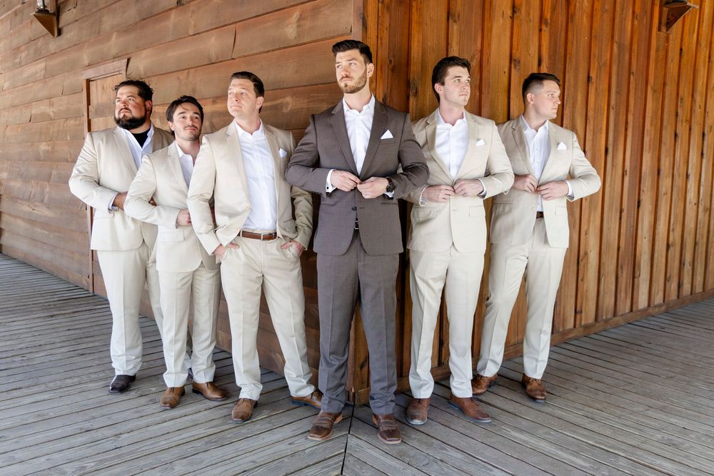 Groom in navy suit stands with groomsmen wearing light beige suits at wooden venue during wedding celebration.