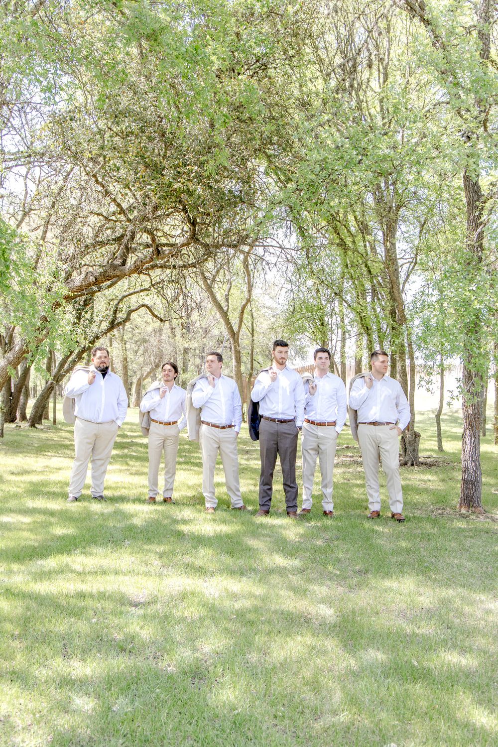 A group gathered under tall trees in a sunlit woodland setting wearing coordinated light colored attire.
